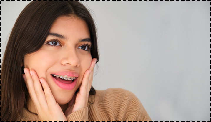 Smiling woman with hands on face wearing tan sweater against neutral background.