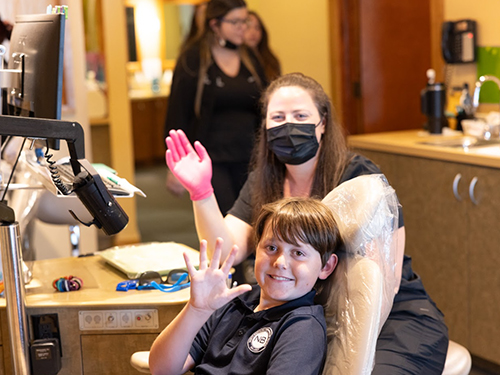 Young child in dental chair waving at camera during orthodontic appointment with masked staff member.