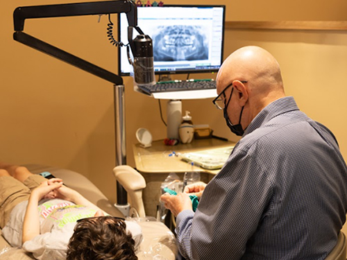 Orthodontist examining patient in dental chair with intraoral camera monitor visible in background.