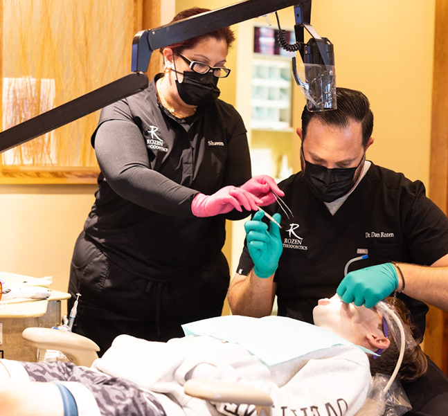 Two orthodontic professionals in masks and gloves examine dental equipment in a clinical treatment room.