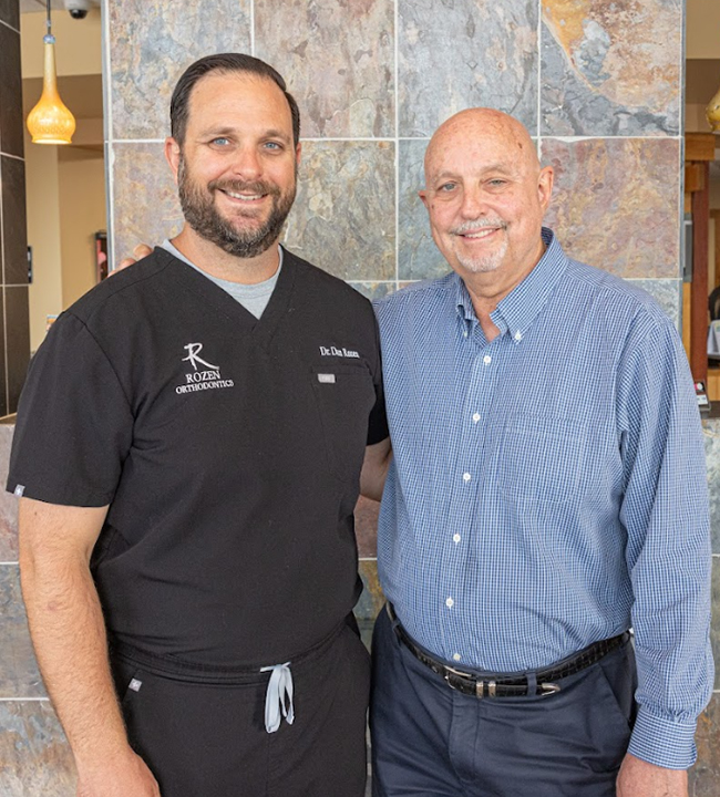 Two men smiling at camera in modern orthodontic office setting with tile wall background.